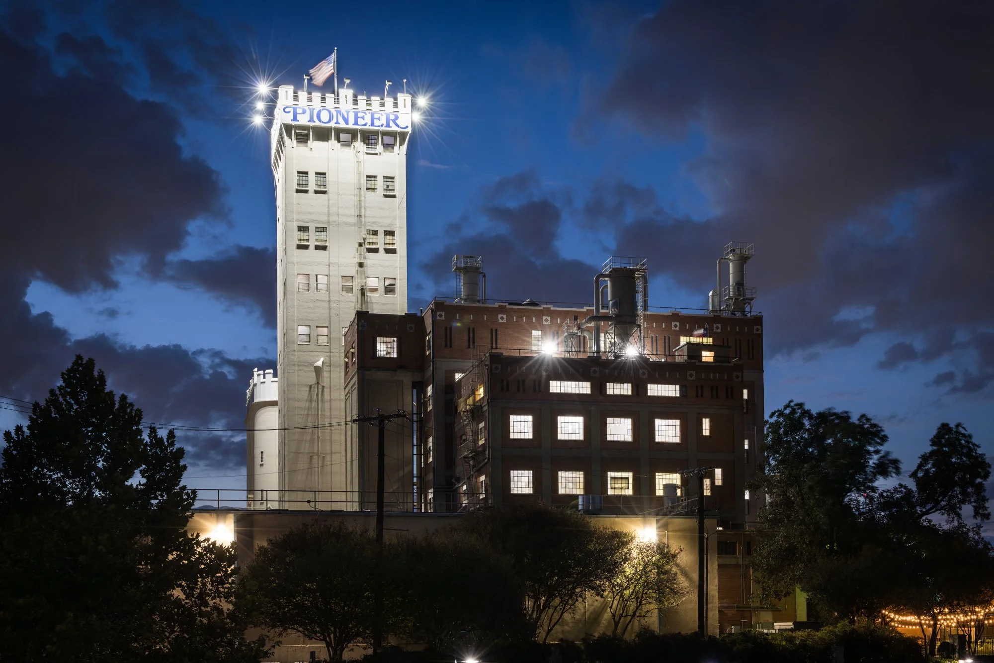 Nighttime view of a tall, illuminated building with the word 'PIONEER' on top, featuring a flag and bright lights, with surrounding trees and dark clouds in the sky.