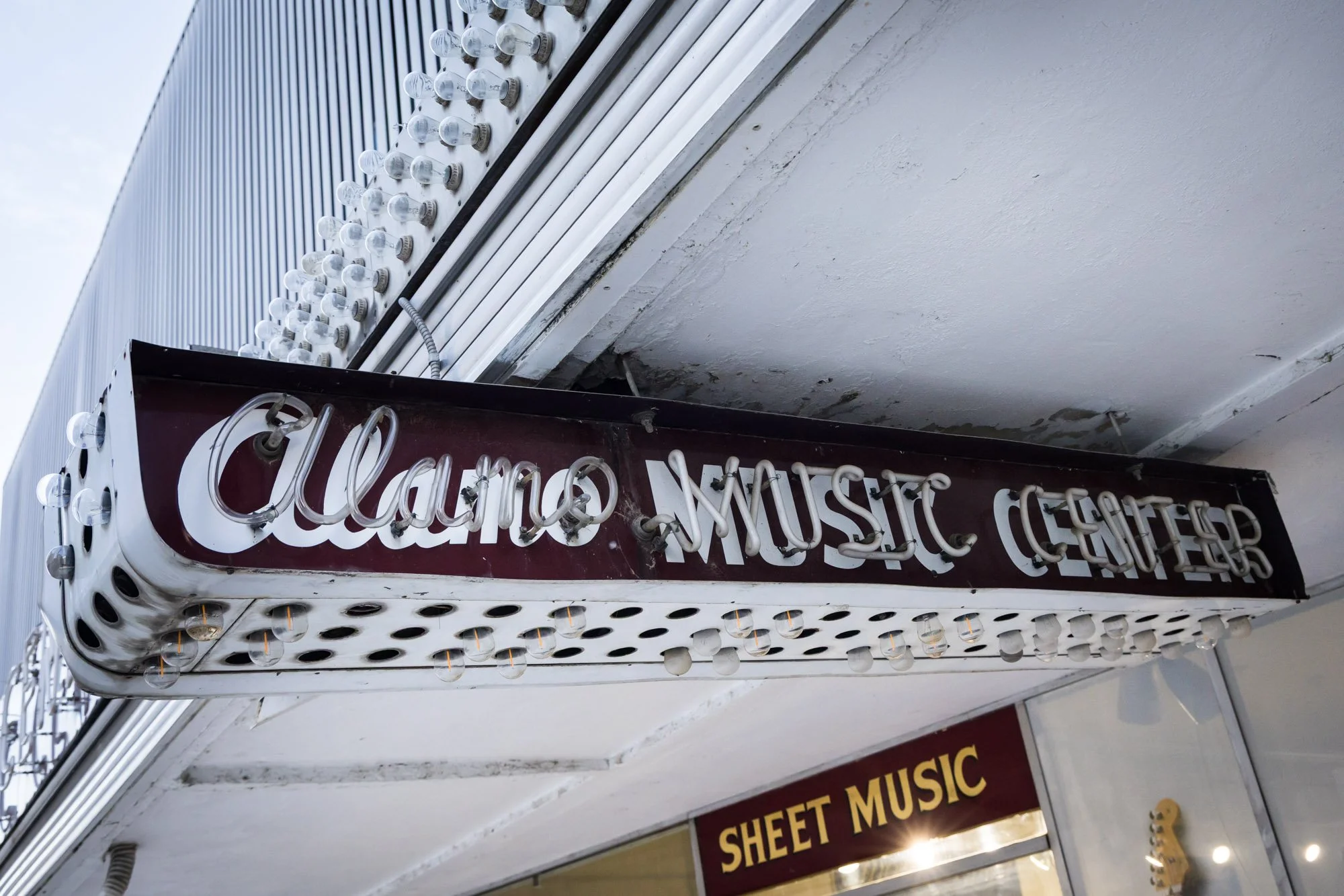 Close-up of a vintage sign for the 'Alamo Music Center' with neon tubing and light bulbs, mounted above a storefront.
