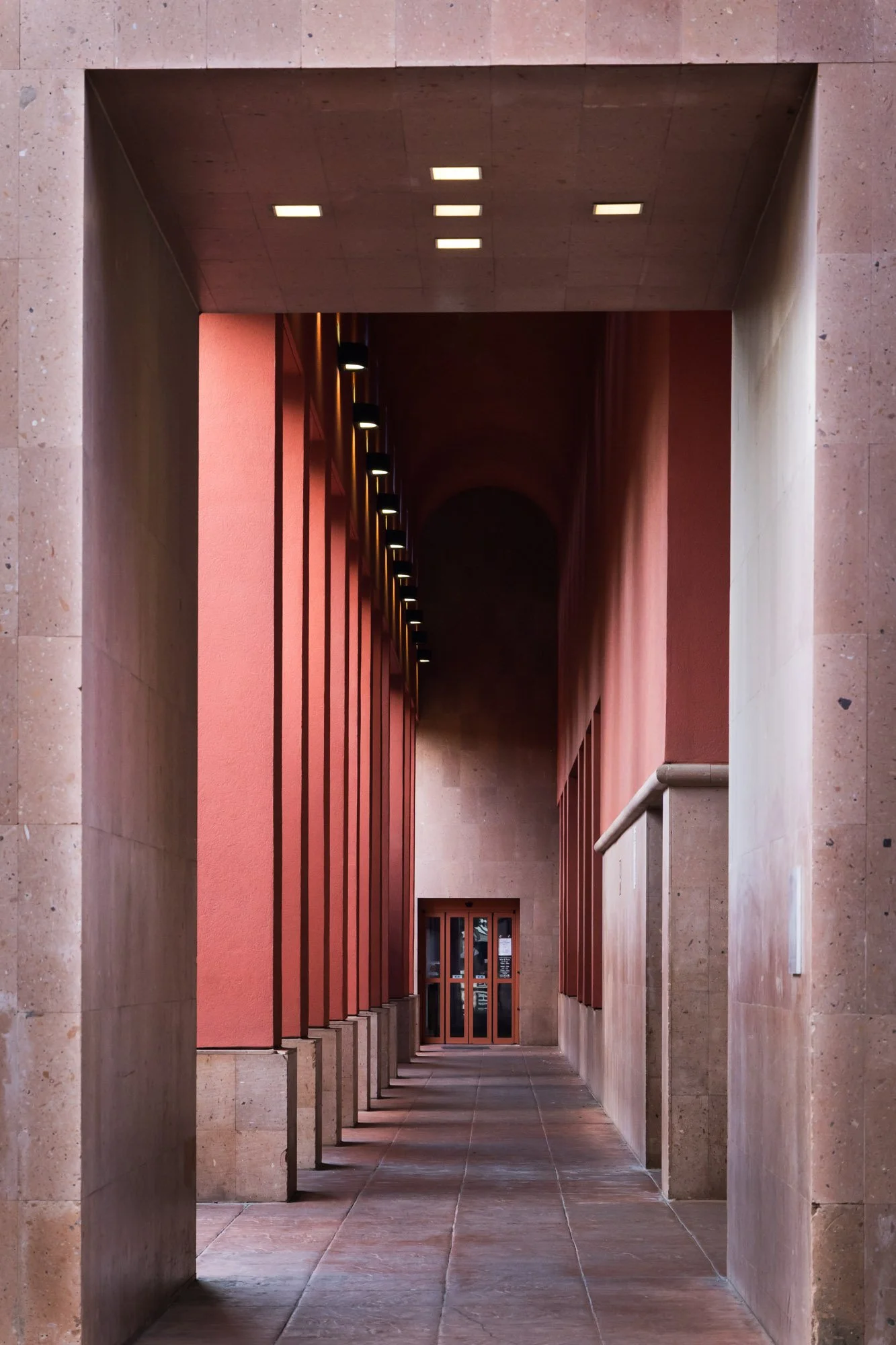 A modern architectural corridor with beige stone walls, a pink wall with vertical wooden slats, and a row of ceiling lights leading to a door at the end.