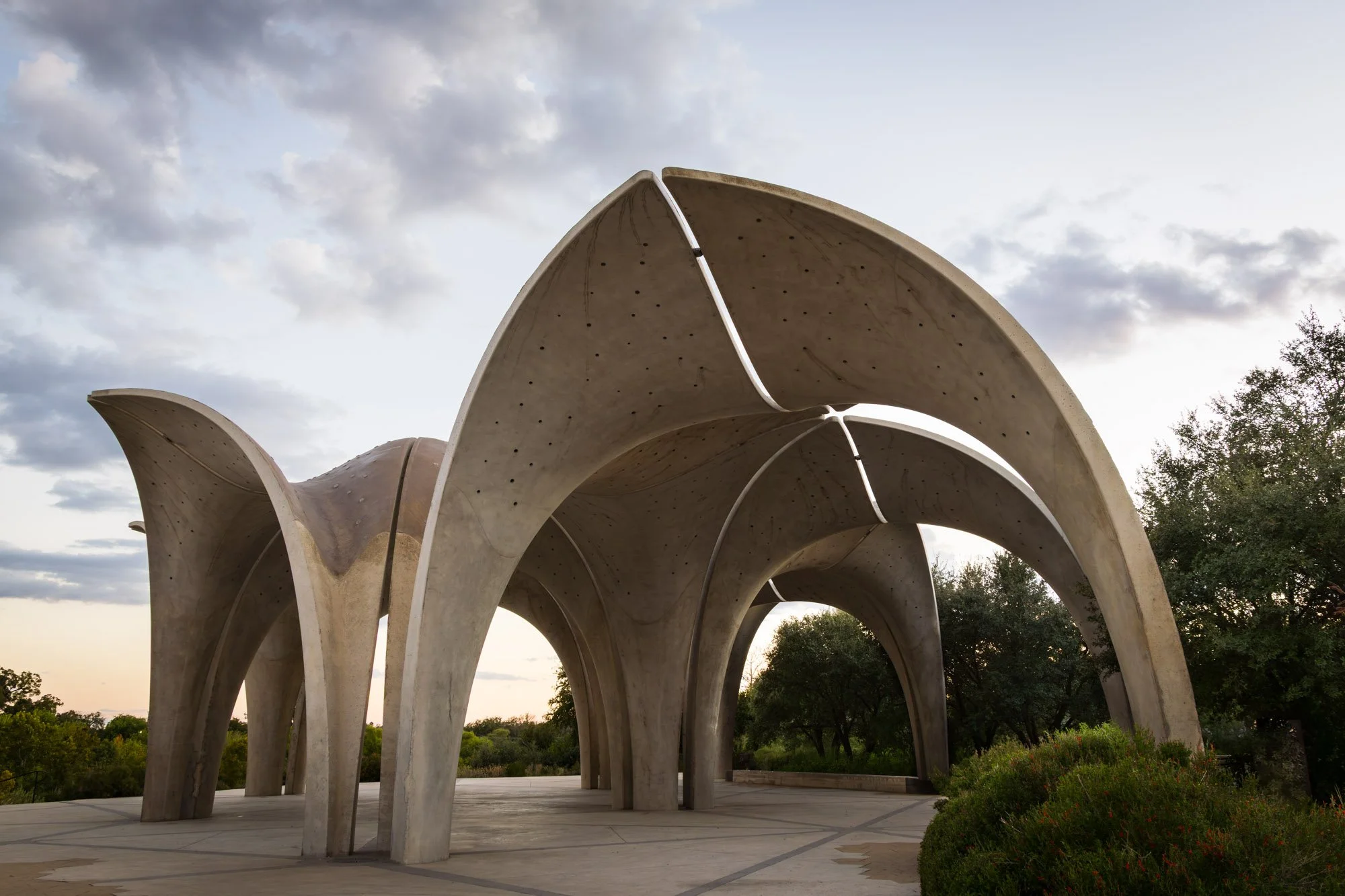 Modern concrete sculpture with large, curved archways outdoors at sunset with trees and cloudy sky in background.