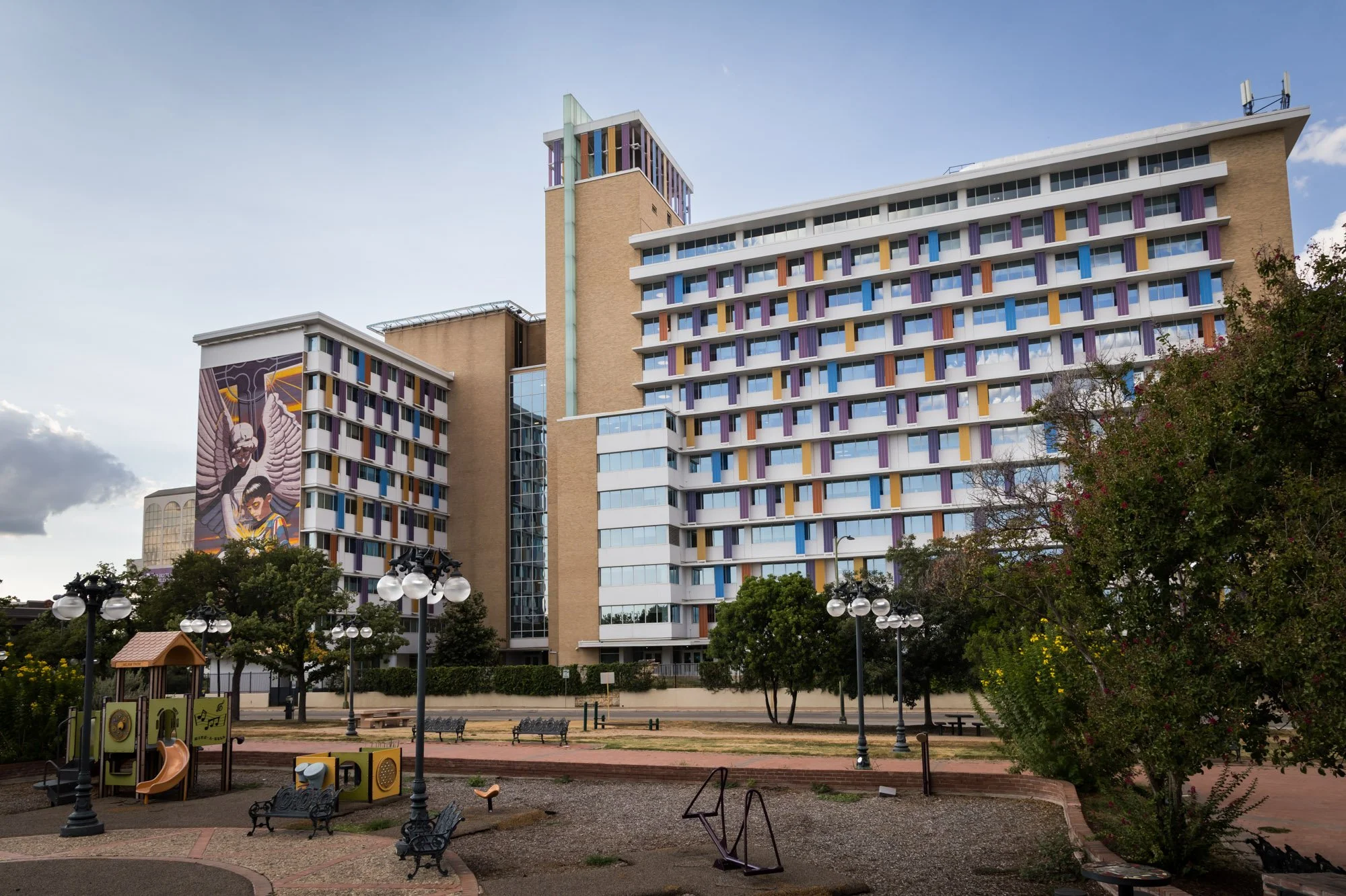 A city park with playground equipment, benches, and lamp posts in front of a large, colorful hotel building with a mural on the side depicting a angel and a person.
