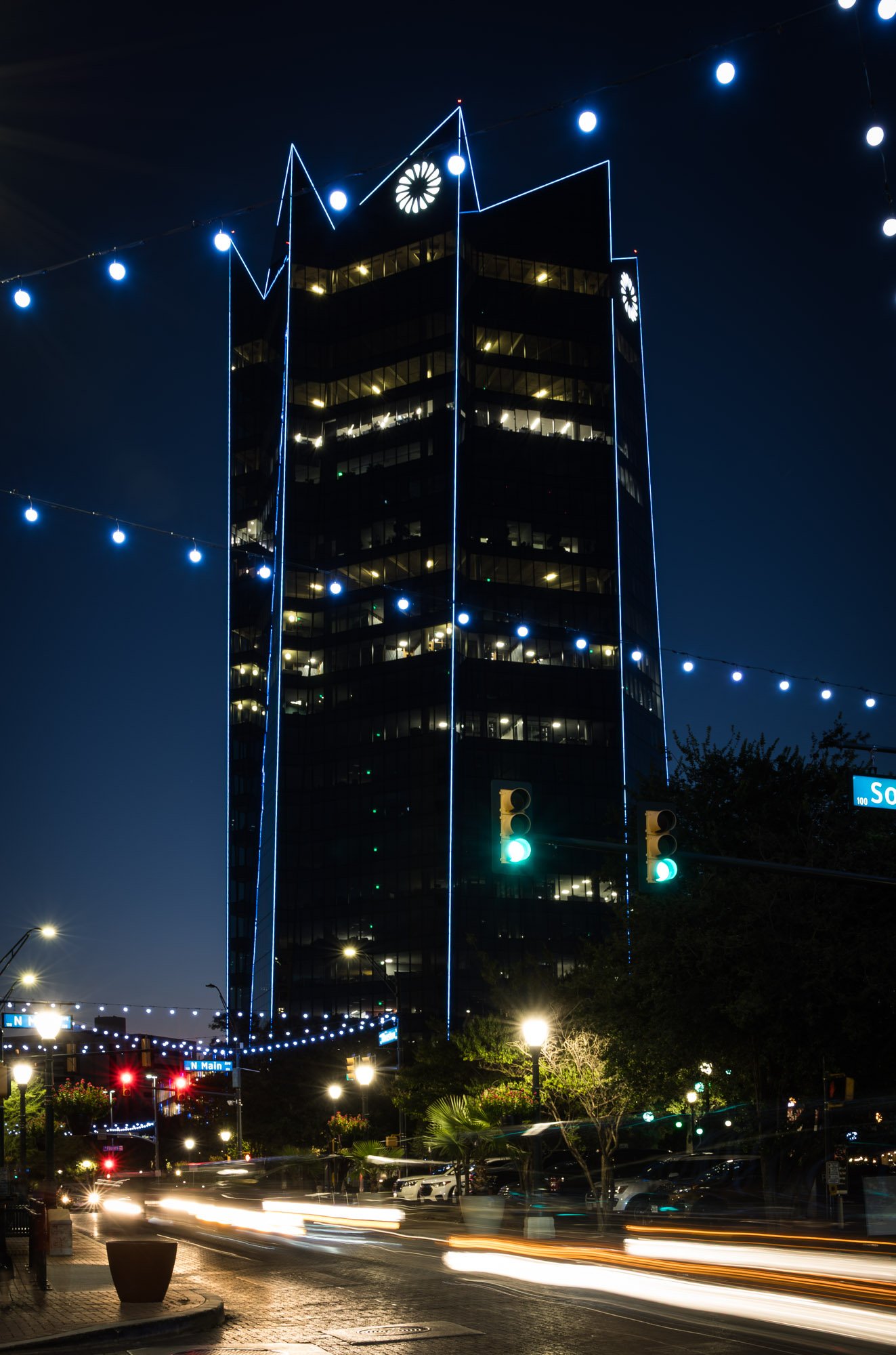 Night scene of a tall glass office building illuminated with blue neon lights outlining its edges, with several windows lit up and decorative lights strung across the street below, along with moving lights from passing cars.