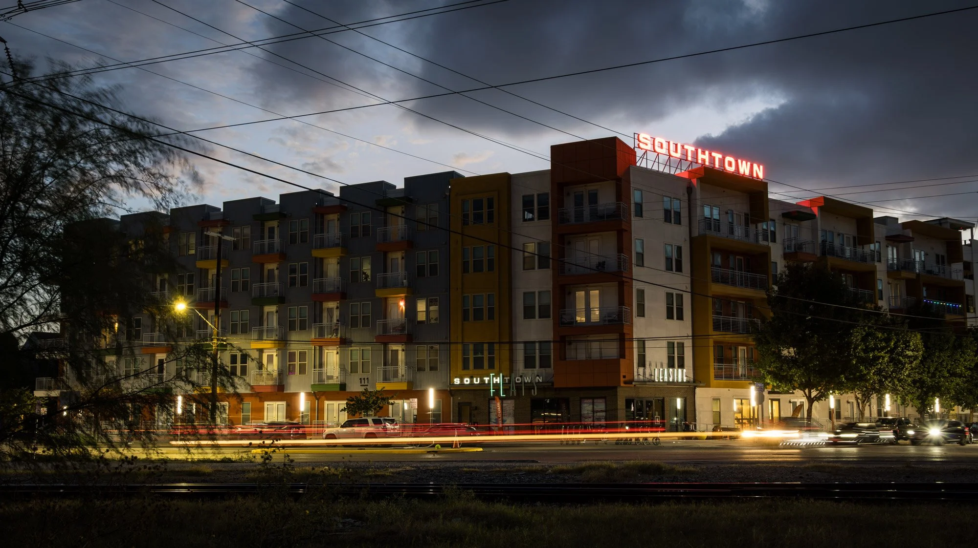A multi-story apartment building with a illuminated sign reading 'SOUTHTOWN' on the roof, nighttime setting, with trees and moving cars in front.
