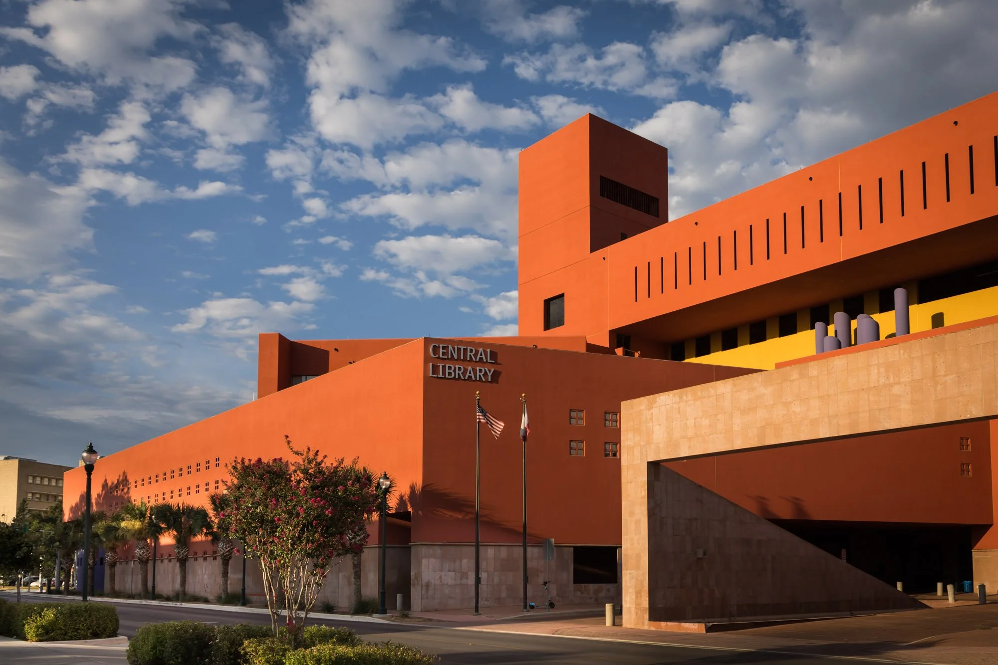 The Central Library building with an orange and yellow facade under a partly cloudy sky, with flags and trees in front.