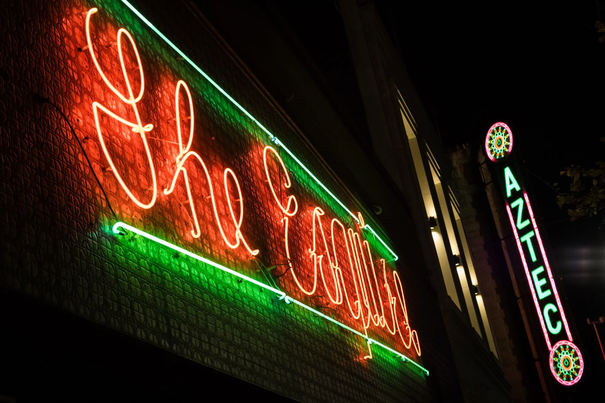 Neon sign with the words "Xhochee" in bright red cursive, outlined in neon green, mounted on a textured wall outside a building at night.