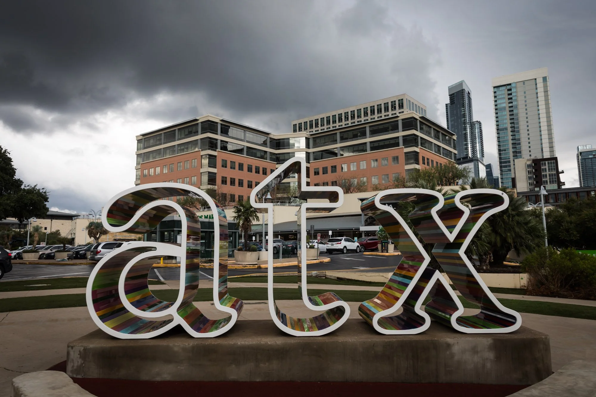 Colorful rounded letters spell out 'austin' in front of a cityscape with modern buildings and a cloudy sky.