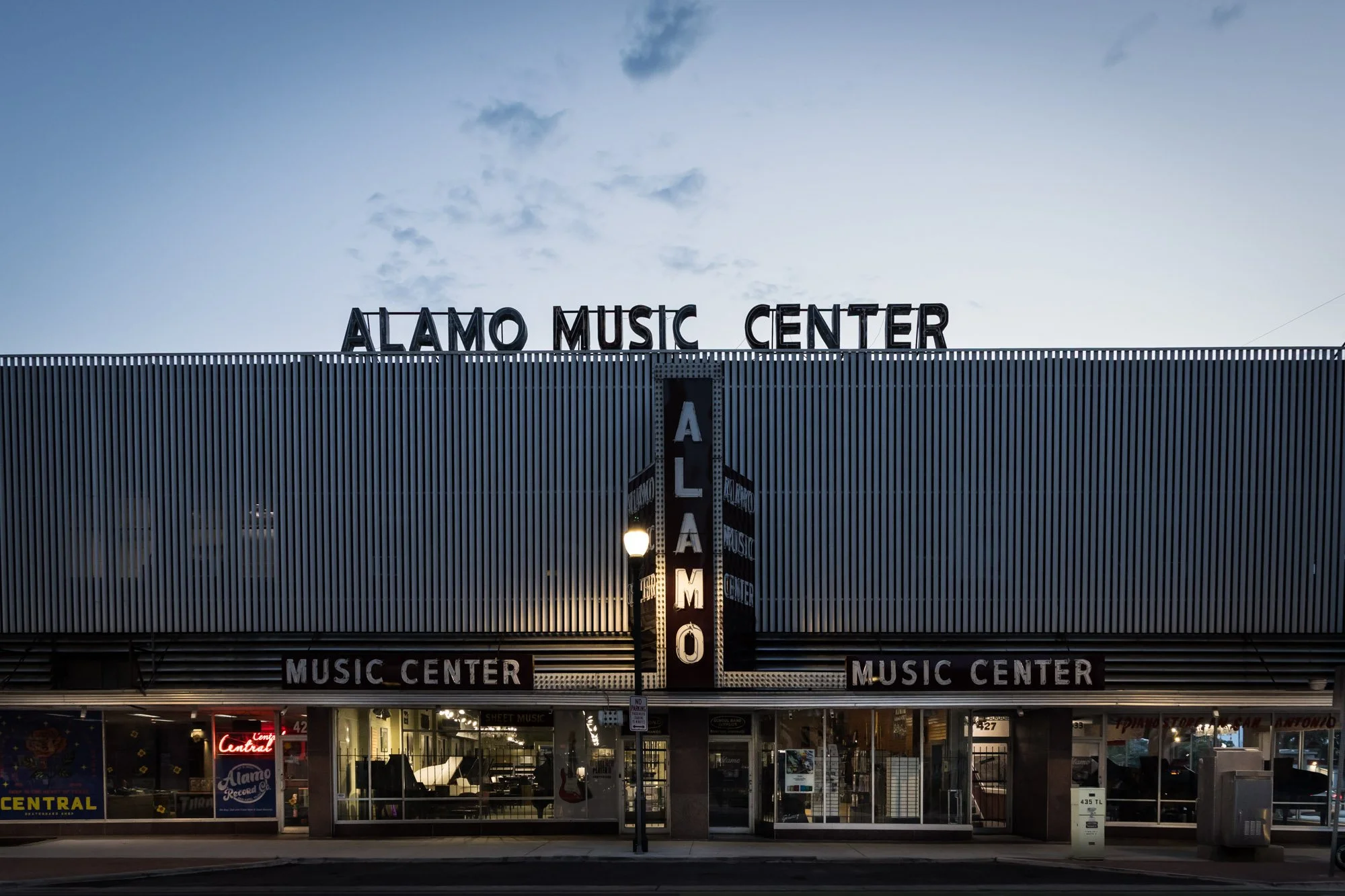 Front view of the Alamo Music Center building at dusk, with signage on the facade and illuminated windows, in an urban setting.