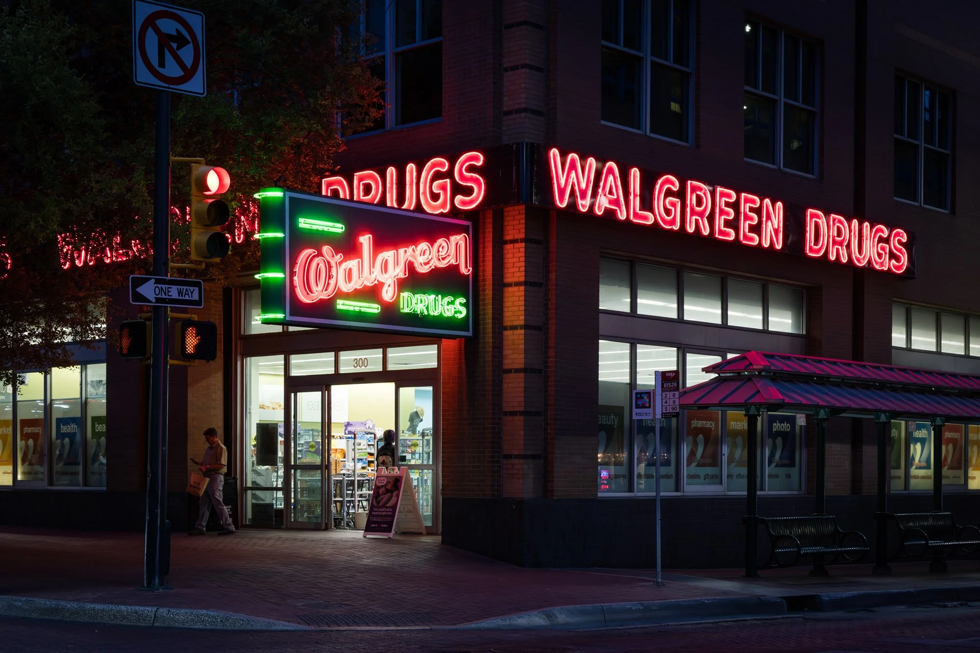 Nighttime exterior view of a Walgreens pharmacy with bright neon signs that read 'Walgreen Drugs' and 'Drugs'. The store has large glass windows, a small awning, and people inside and outside the entrance.