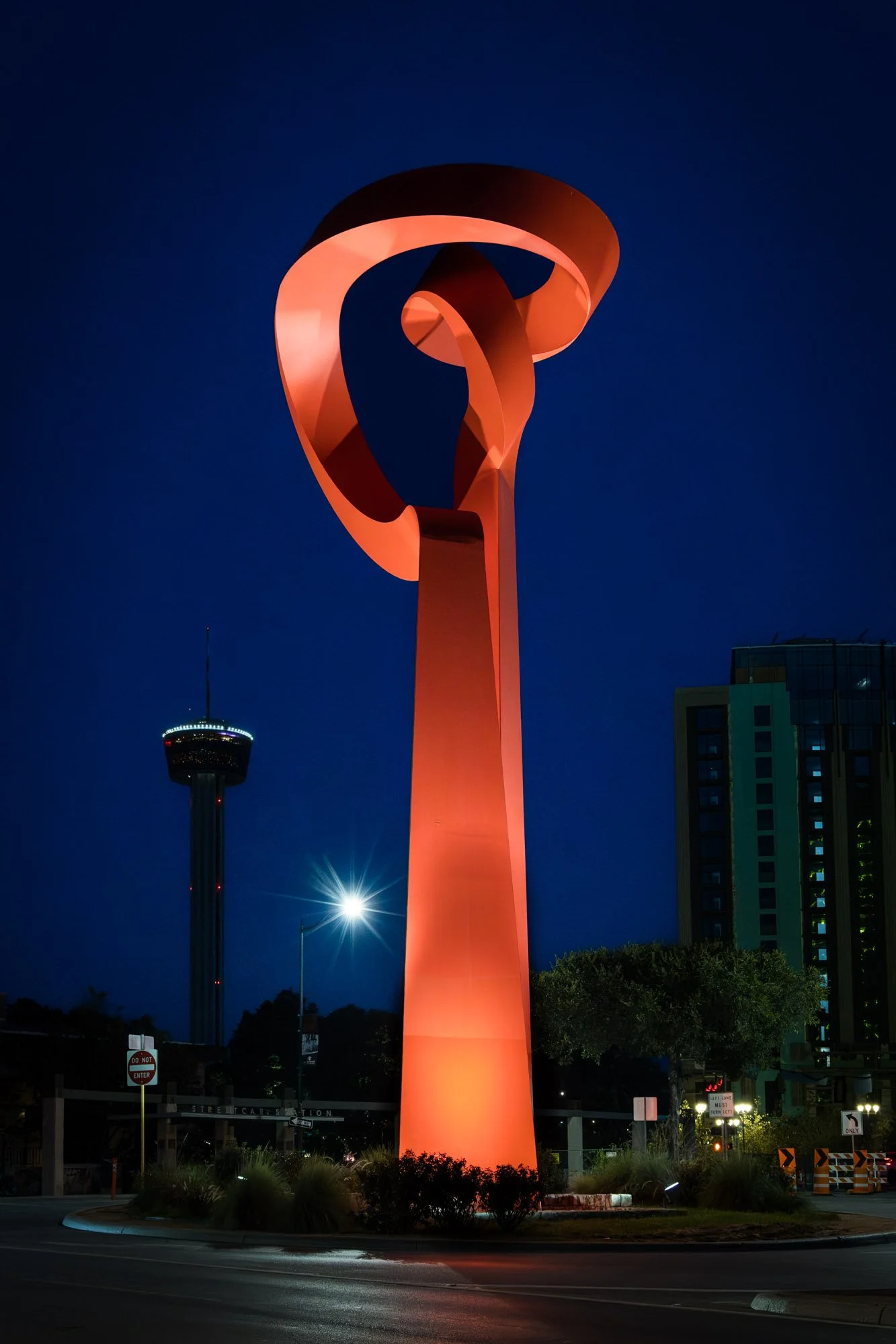 Nighttime view of a tall, abstract sculpture with swirling, looped design illuminated in orange, with a city building and the Needle in the background.
