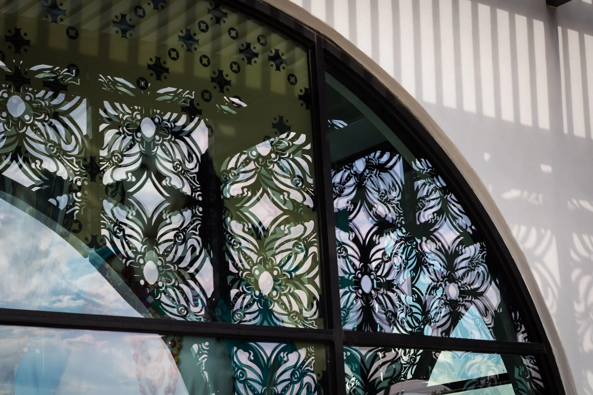 Decorative black metal window grill with intricate floral patterns, reflecting outside sky and clouds, set in a white arched wall.