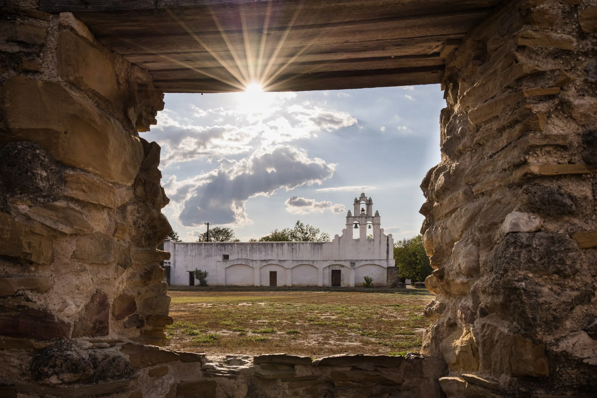 View of a white historic church through a stone window opening with the sun shining behind the roof, surrounding trees, and cloudy sky in the background.