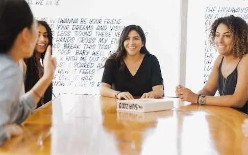 Three women sitting around a wooden table engaged in conversation, with whiteboards with handwritten notes and lyrics in the background.