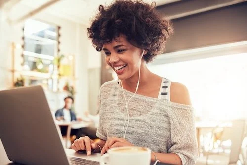 Young woman with curly hair smiling and wearing headphones while working on a laptop in a cozy cafe