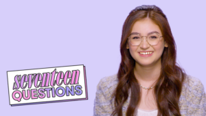 A young woman with long brown hair and glasses smiling against a light purple background, next to a sign that reads "seventeen QUESTIONS" in purple and pink letters.