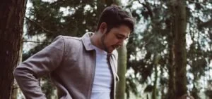 Man wearing a gray blazer looking down outdoors with trees in the background.