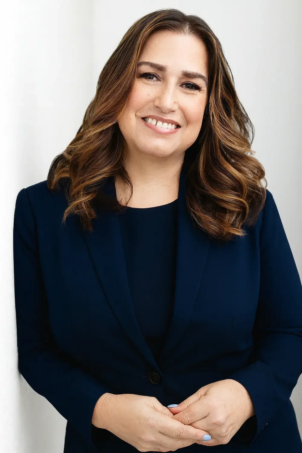Portrait of a woman with brown hair, smiling, wearing a dark blue blazer and matching top, standing against a white background.