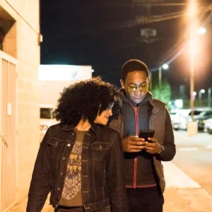 Two people walking on a city street at night, looking at a smartphone, illuminated by streetlights.