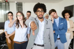 A group of diverse professionals in business casual attire standing together indoors, with one man in front giving a thumbs down.