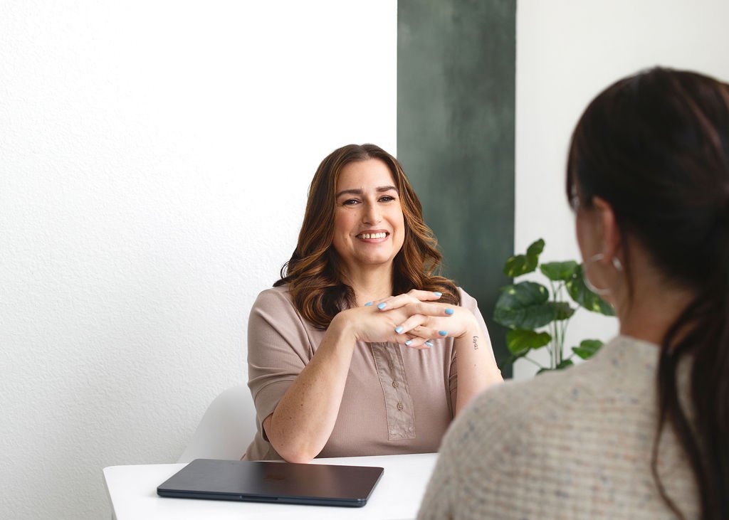 Two women having a conversation in an office, one visible with brown hair, wearing a beige shirt, smiling, and the other with dark hair, wearing glasses and a beige sweater.