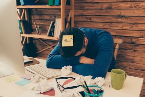 A man sitting at a cluttered desk with crumpled papers, a green mug, and eyeglasses, resting his head on his arms with a note reading 'HELP' on his baseball cap.