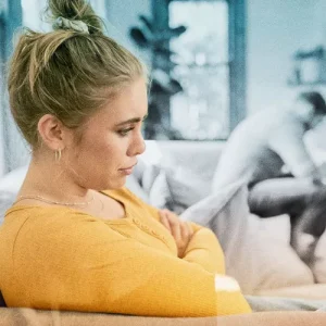 A woman with blonde hair in a bun wearing a yellow sweater sitting on a couch, looking down.