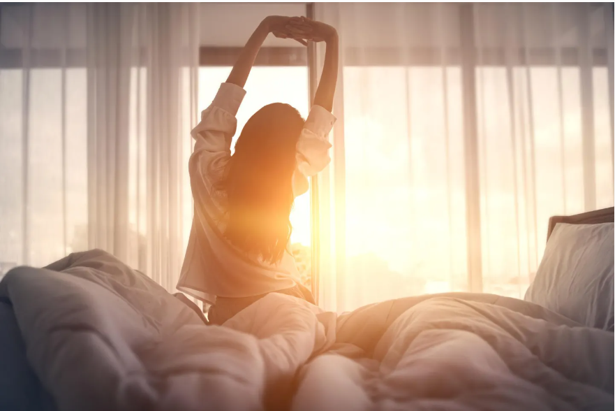 A woman stretching in bed during sunrise, sunlight coming through curtains.