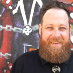 A bearded man smiling in front of a graffiti mural.