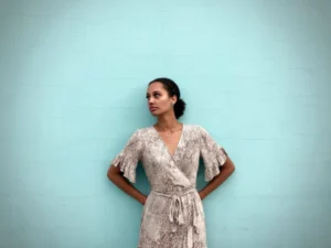 Woman in a light-colored dress standing against a turquoise wall with her hands behind her back