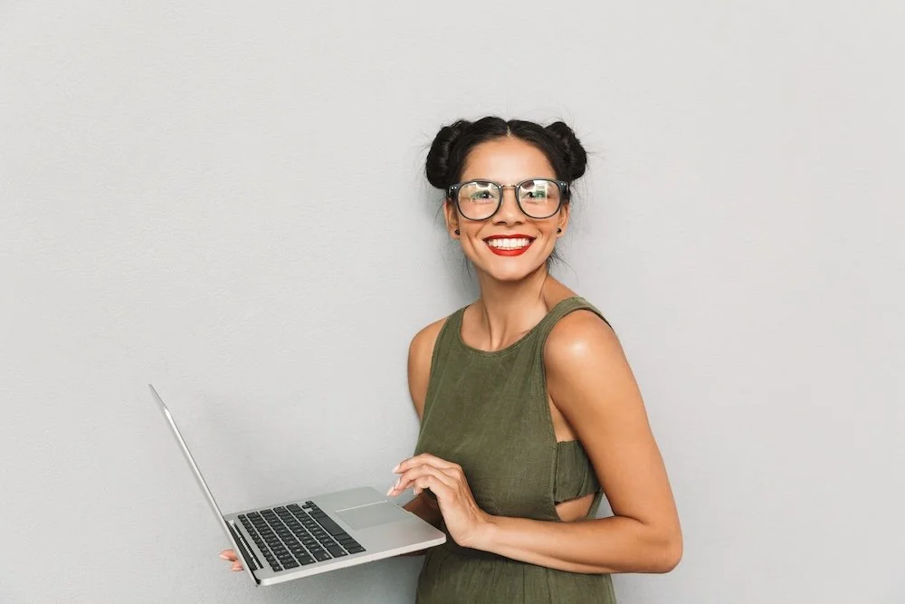 Smiling woman with glasses holding a laptop against a plain wall.