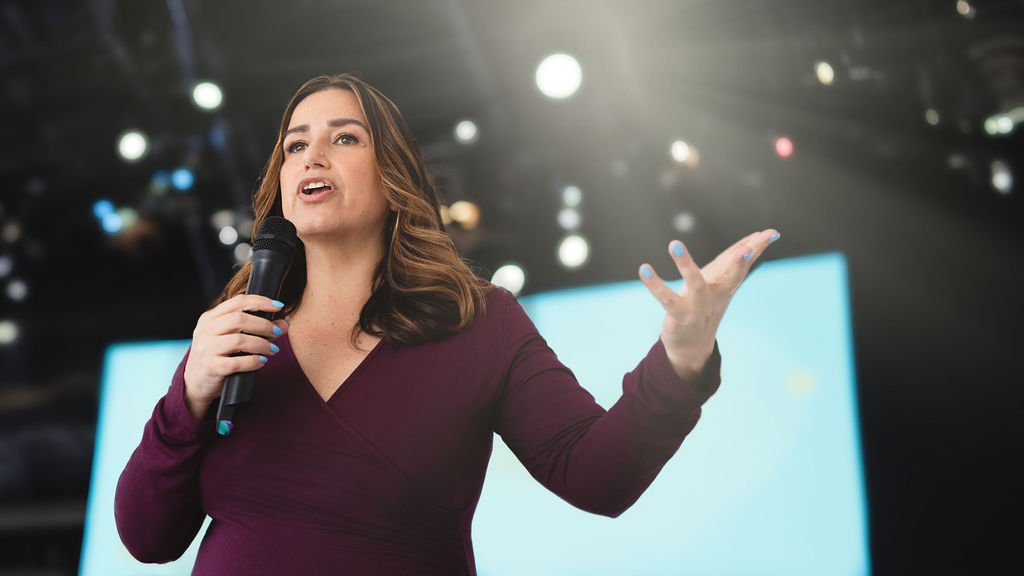 A woman with brown hair holding a microphone and speaking.