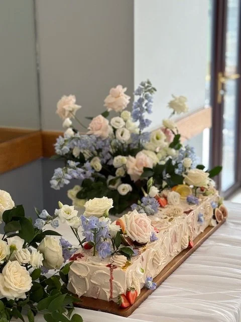 A floral cake decorated with pastel roses and other flowers, with a flower arrangement on top, placed on a table near a window.