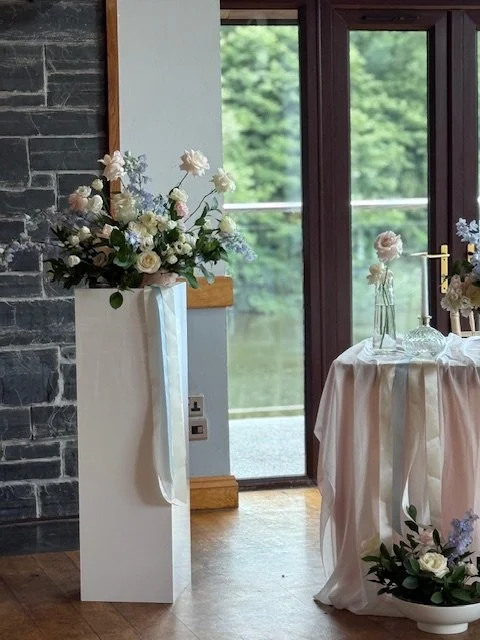 Vase of white and pink flowers on a tall white pedestal near glass door with greenery outside.