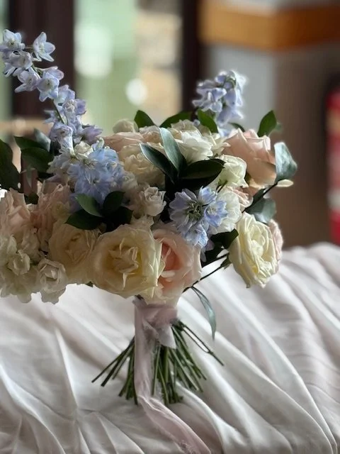 A bouquet of white and pale pink roses with blue and purple flowers, tied with a pink ribbon, resting on a white fabric surface.
