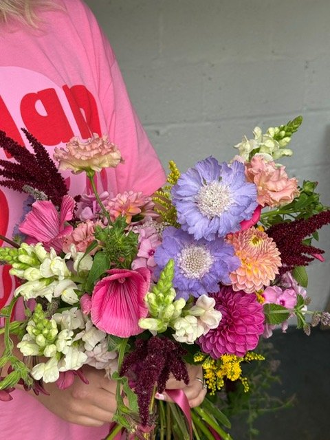Person in a pink shirt holding a colorful bouquet of mixed flowers, including pink, purple, white, and yellow blossoms.