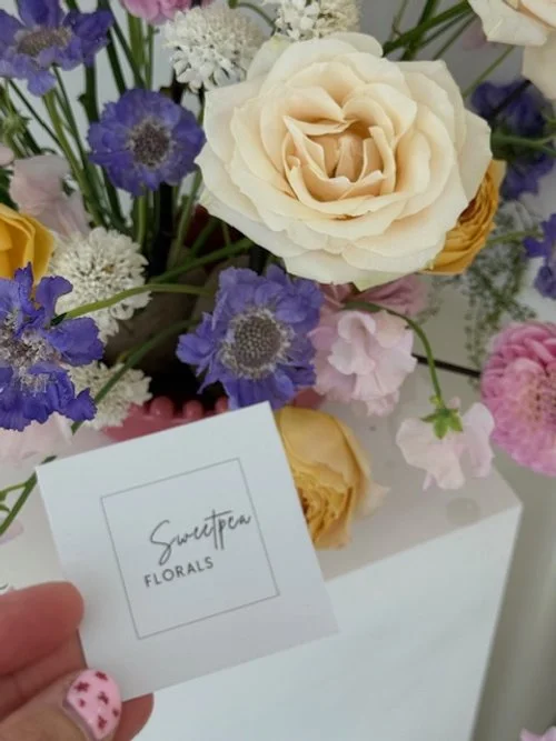 Mixed bouquet of white, purple, pink, and peach flowers with a business card reading 'Sweetpea Florals' in front.