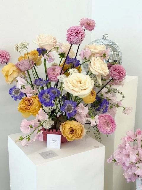 A colorful floral arrangement with roses, carnations, and other flowers in pastel yellow, pink, purple, and cream, placed on a white pedestal, with a small card and a disco ball decoration in the background.