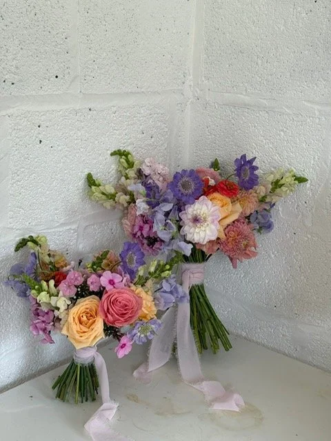 Two bouquets of mixed pastel and purple flowers with white ribbon on a white surface against a white brick wall