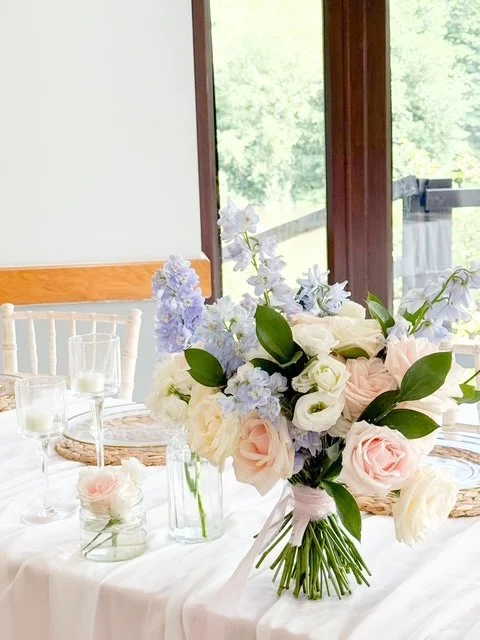 Elegant floral centerpiece with pink roses, white roses, and light purple flowers on a table set for a special occasion.