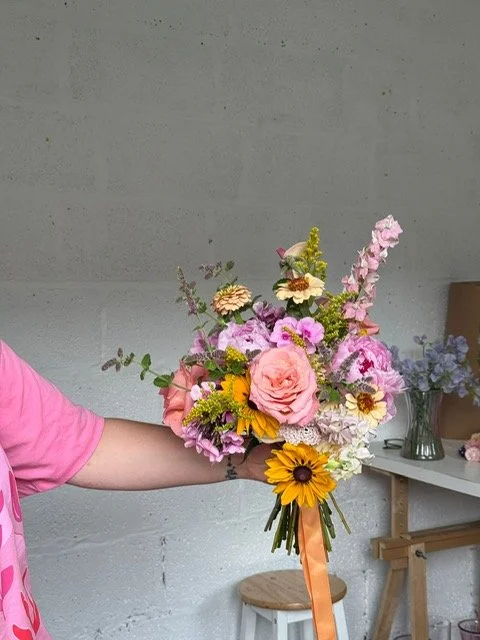 Hand holding a colorful bouquet of flowers including pink roses, yellow sunflowers, and various pink and purple blossoms