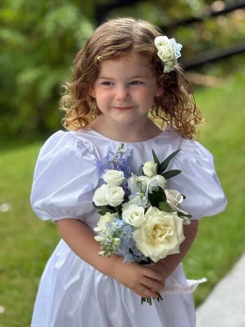 Young girl with curly hair wearing a white dress and floral headpiece, holding a bouquet of white and light purple flowers outdoors.