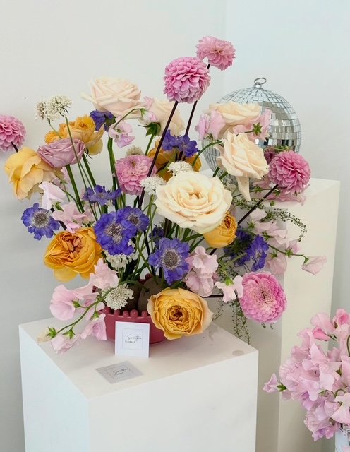A colorful bouquet of various flowers, including roses, dahlias, and anemones, arranged on a white pedestal with a small informational card. A silver disco ball is visible in the background.
