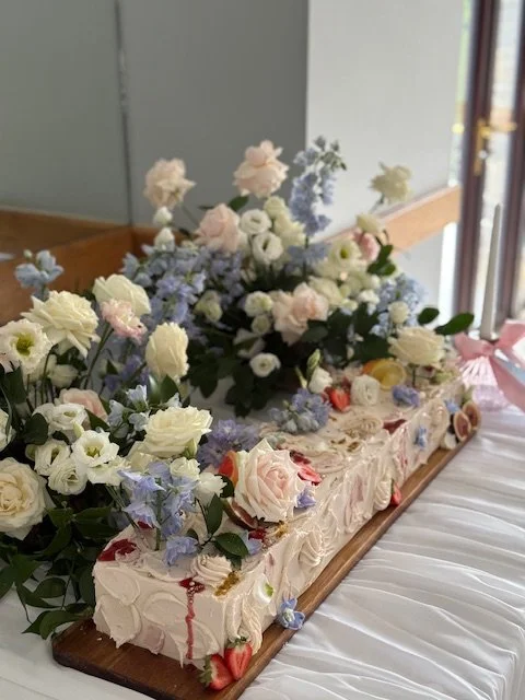 Decorative cake with white frosting topped with an arrangement of white and pastel-colored flowers, including roses and other blooms, on a wooden board.