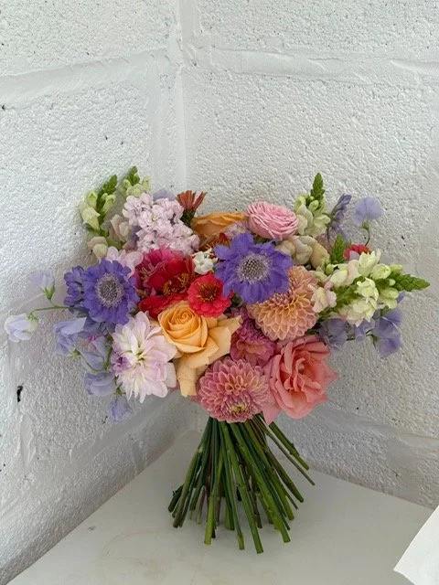 Colorful mixed flower bouquet with roses, dahlias, and other blossoms in pink, purple, orange, and white, arranged in a glass vase on a white surface against a white brick wall.