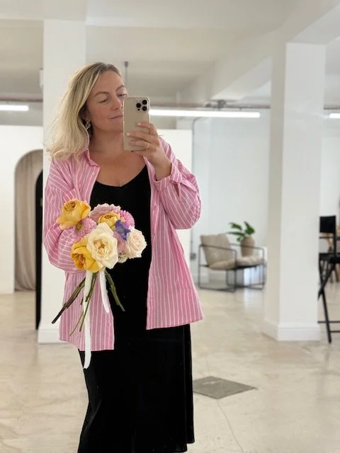 Woman taking a mirror selfie holding a bouquet of colorful flowers, wearing a black dress and a pink-striped shirt in a bright, modern room.