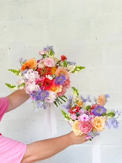 Two hands holding colorful flower bouquets against a white wall.