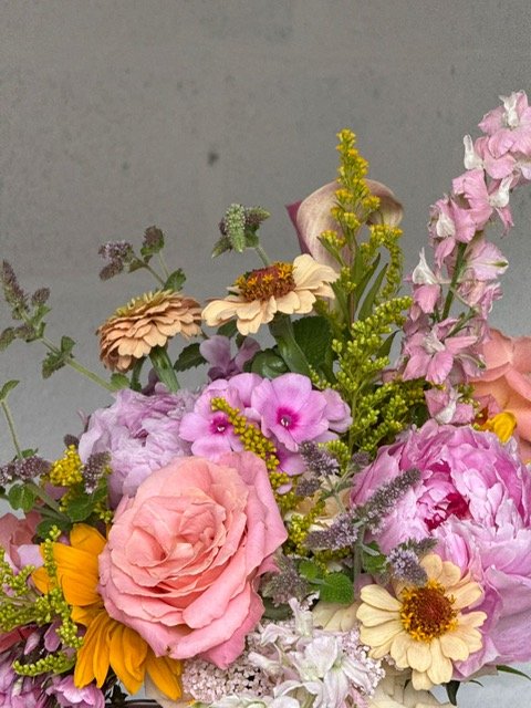 Close-up of a colorful flower bouquet with pink, peach, purple, yellow, and white flowers, including roses and daisies, against a plain background.