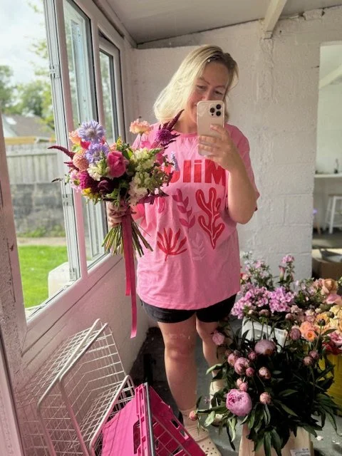 Woman taking a mirror selfie with a bouquet of colorful flowers in a bright room with large windows, floral arrangements on the floor, all in a cheerful setting.