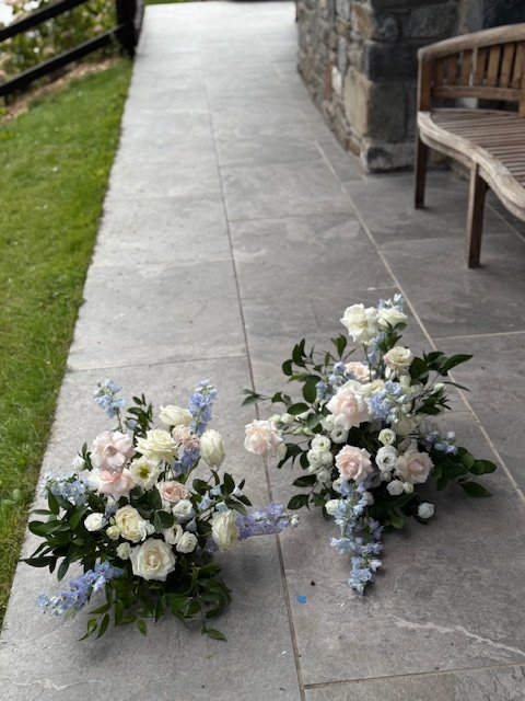 Two floral arrangements with white and light purple flowers on a stone patio near a wooden bench.