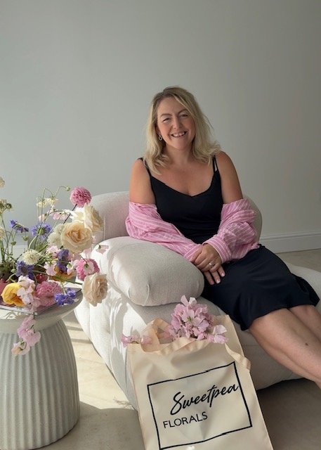A woman sitting on a white sofa, smiling, next to a large bouquet of colorful flowers, with a shopping bag labeled 'Sweetpea Florals' on the floor.