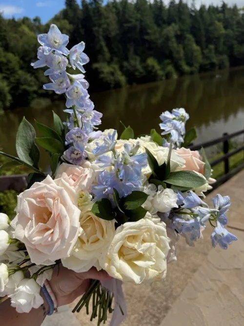 A bouquet of white and light pink roses and blue delphinium flowers being held outdoors near a river with trees in the background.